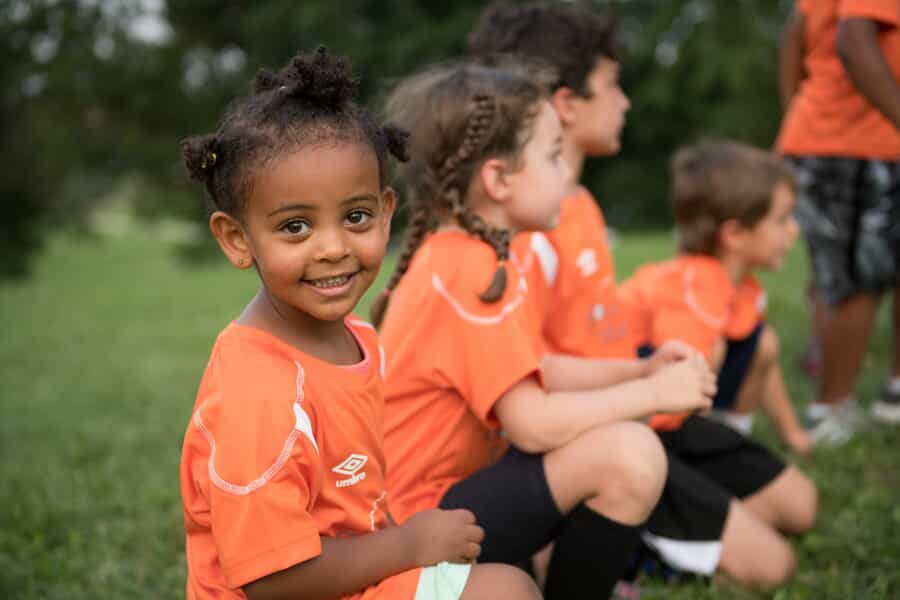 young kids sit on a soccer field wearing orange uniforms